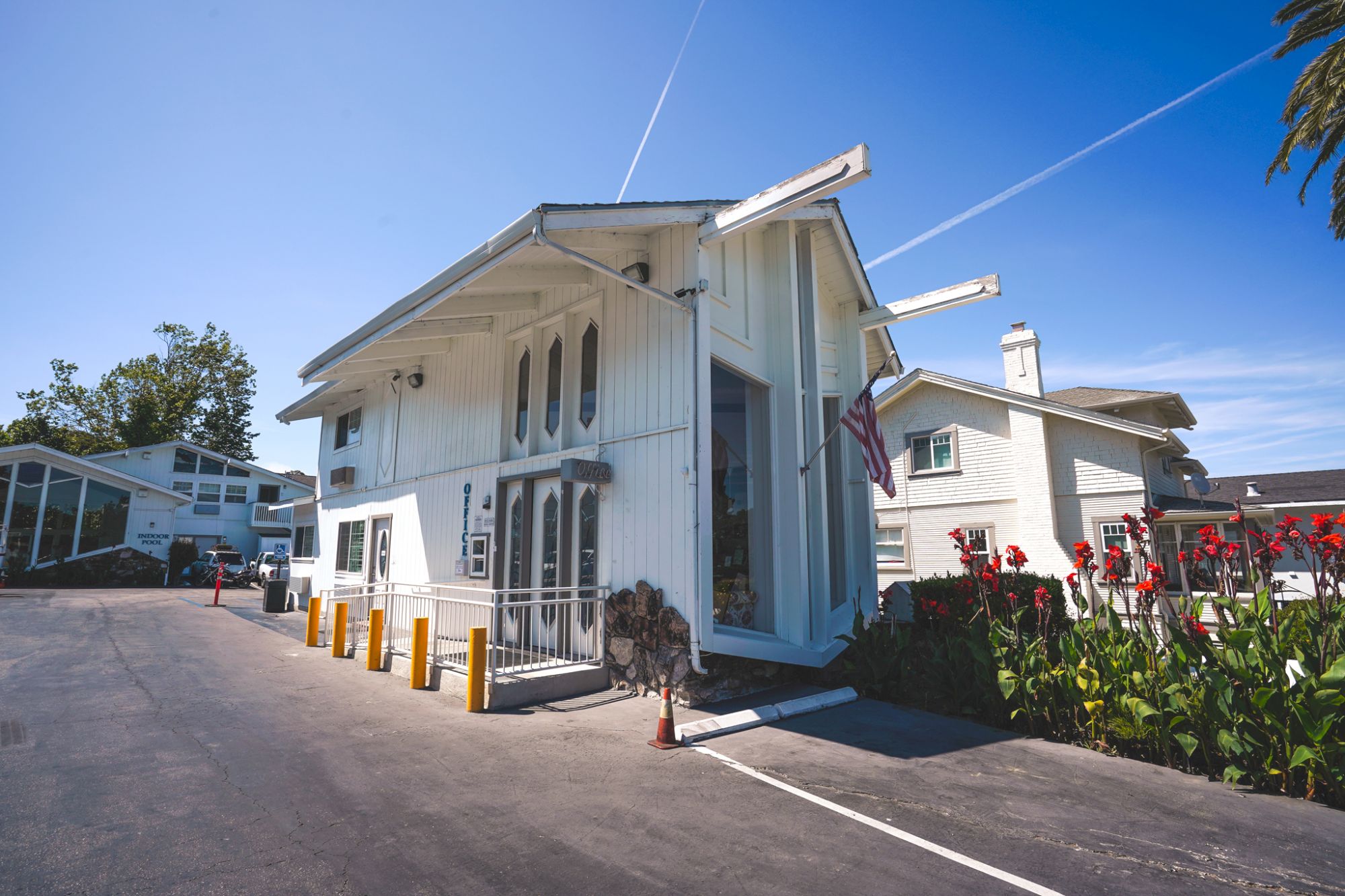 White modern building with tall glass walls, blue sky, palm trees, and a small parking lot; looks like a bright, airy clubhouse or cafe.