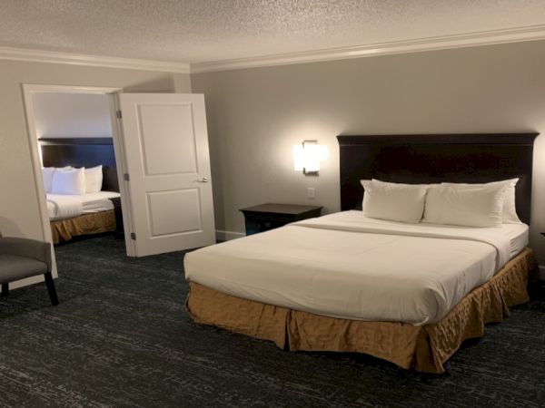 The image shows a hotel room with a bed, two nightstands, a chair, and an open door leading to another room with another bed.