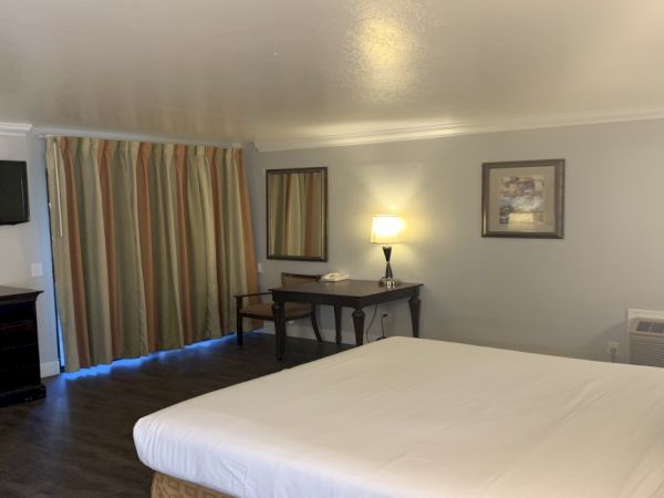 The image shows a tidy hotel room with a bed, wall-mounted TV, desk, lamp, curtains, and a framed picture on the wall.
