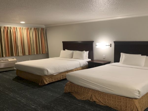The image shows a hotel room with two double beds, a window with curtains, a lamp, and a small table.