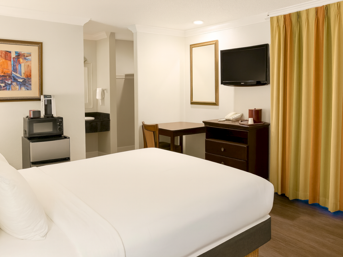 A hotel room with a white bed, a desk, dresser, wall art, a wall-mounted TV, and gold curtains, plus a small microwave area, all in a cozy setup.