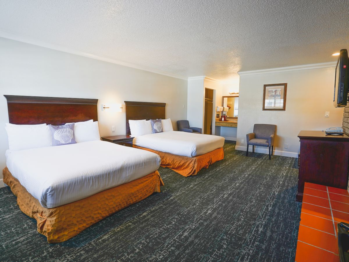 A hotel room with two queen beds, nightstands, a seating area, a desk, a brick fireplace, and a doorway to a small kitchenette.