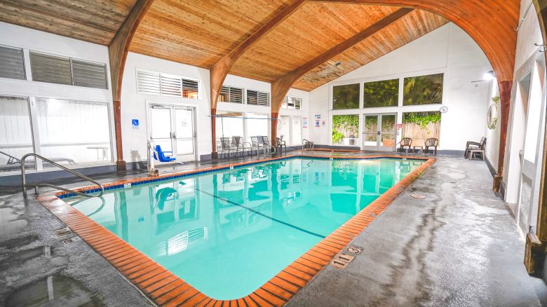Indoor rectangular swimming pool with blue water, brick coping, wooden vaulted ceiling, wet concrete floor, lounge chairs, and large windows.