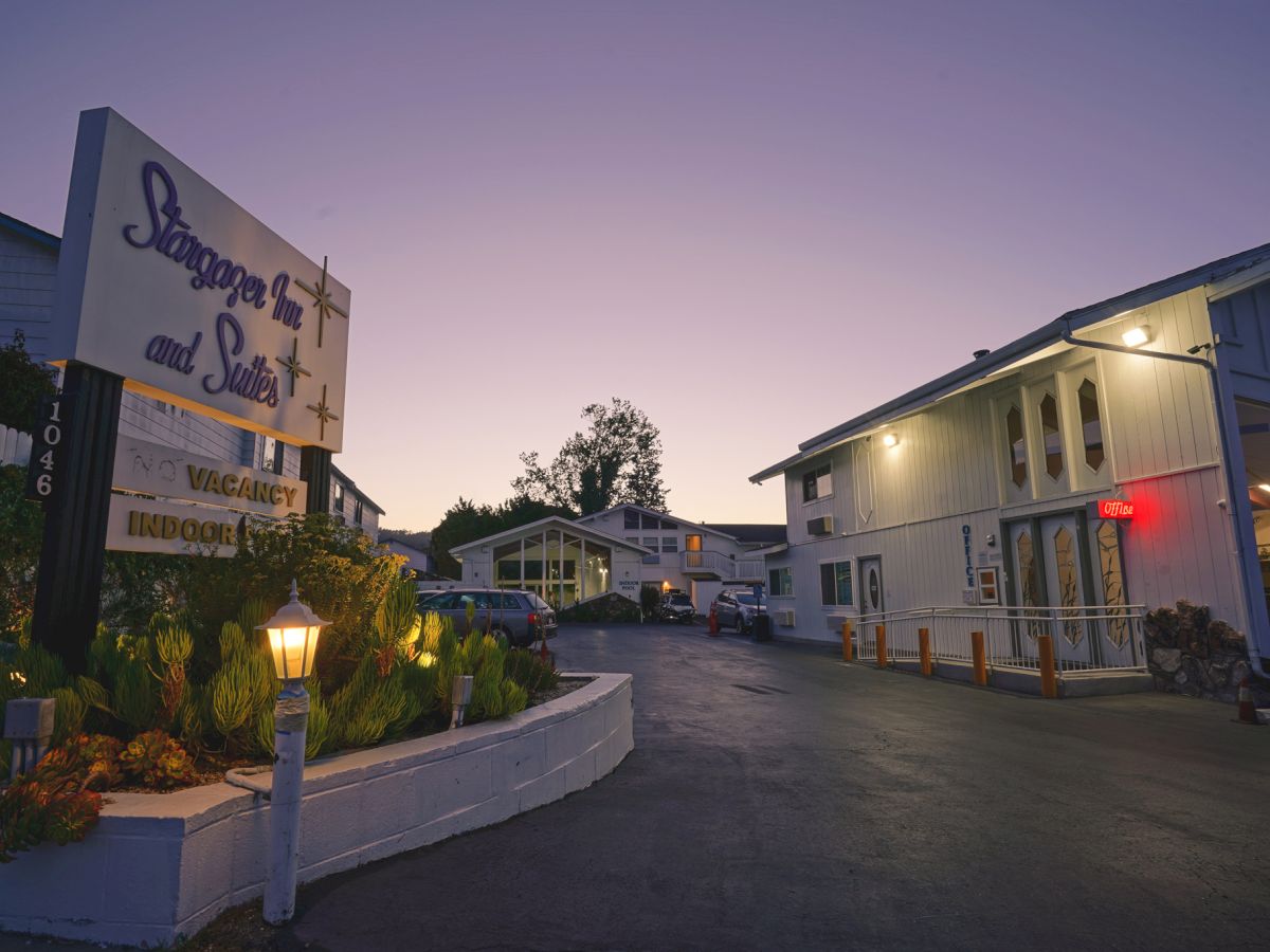 A small motel at dusk with a sign, courtyard, and two- to three-story buildings, dim streetlights, and a quiet, empty driveway.