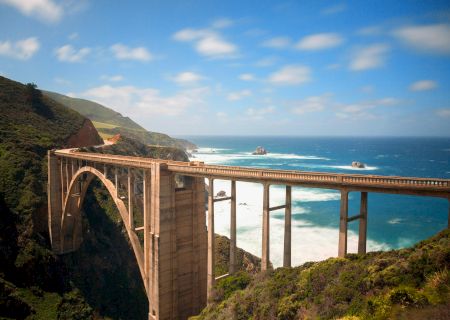 A coastal cliffside view with a stone balcony, railings, blue ocean, and clear sky.