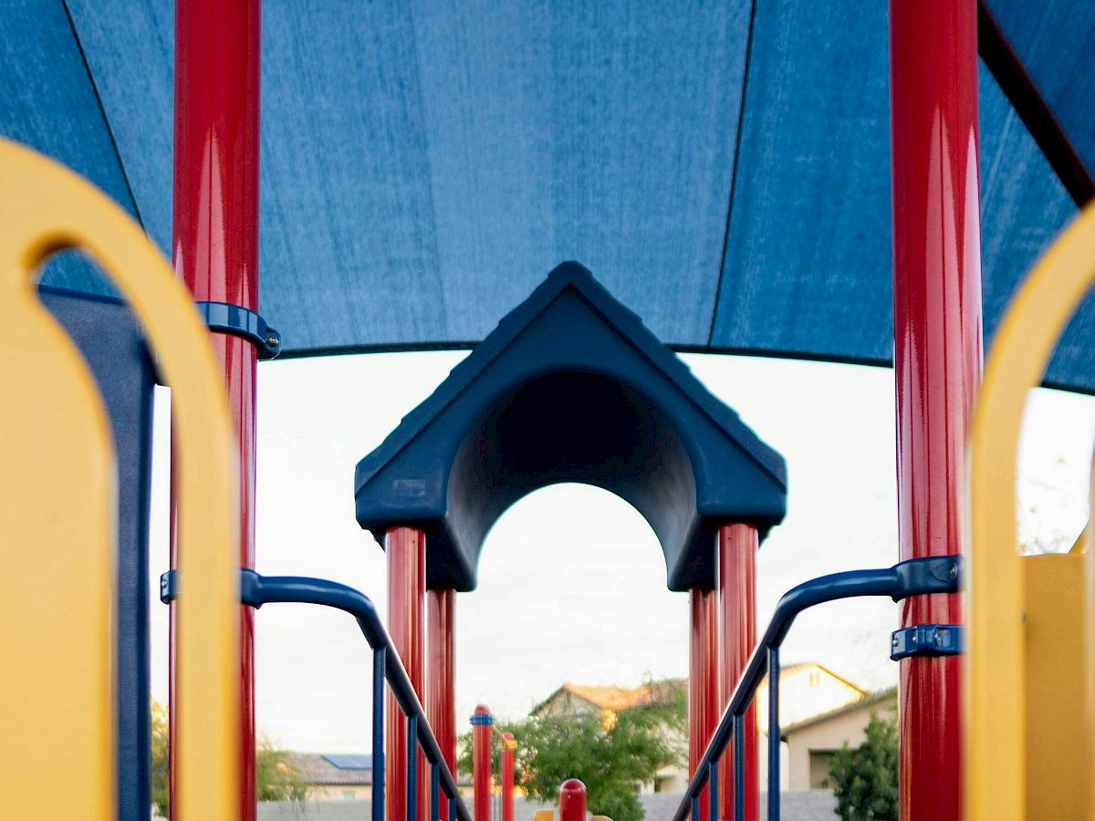 The image shows a playground structure with blue shade, red poles, yellow panels, and platforms for children to play on.