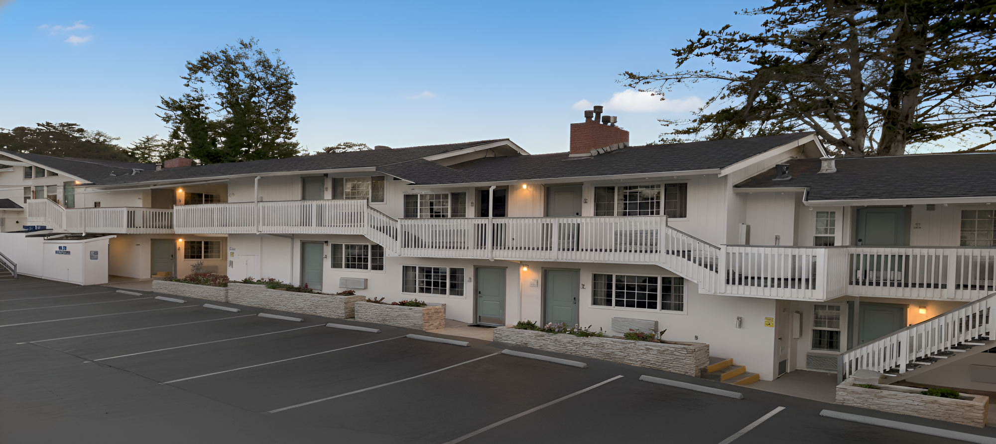 A two-story apartment building with white siding, exterior walkways, staircases, and a parking lot in front, under a clear sky.