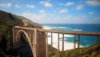 A tall coastal bridge arches over rugged cliffs toward the blue ocean, with green hills, rocky coastline, and a sunny, partly cloudy sky.