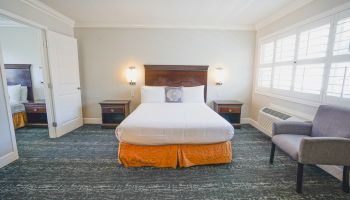 A tidy hotel room with a queen bed, two nightstands, wall lamps, a windowed wall, a chair, and a full-length mirror reflection, neat and bright.