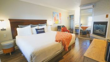 A modern hotel room with a large bed, white linens, orange throw, two nightstands, a seating area, a desk, a TV, and a fireplace near a bright entryway.