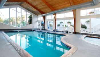 Indoor swimming pool with clear blue water, brick border, wooden ceiling, large windows, and lounge chairs along the far wall, bright and inviting.