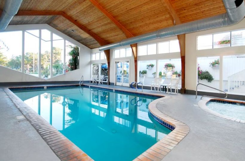 Indoor swimming pool with clear blue water, brick border, wooden ceiling, large windows, and lounge chairs along the far wall, bright and inviting.