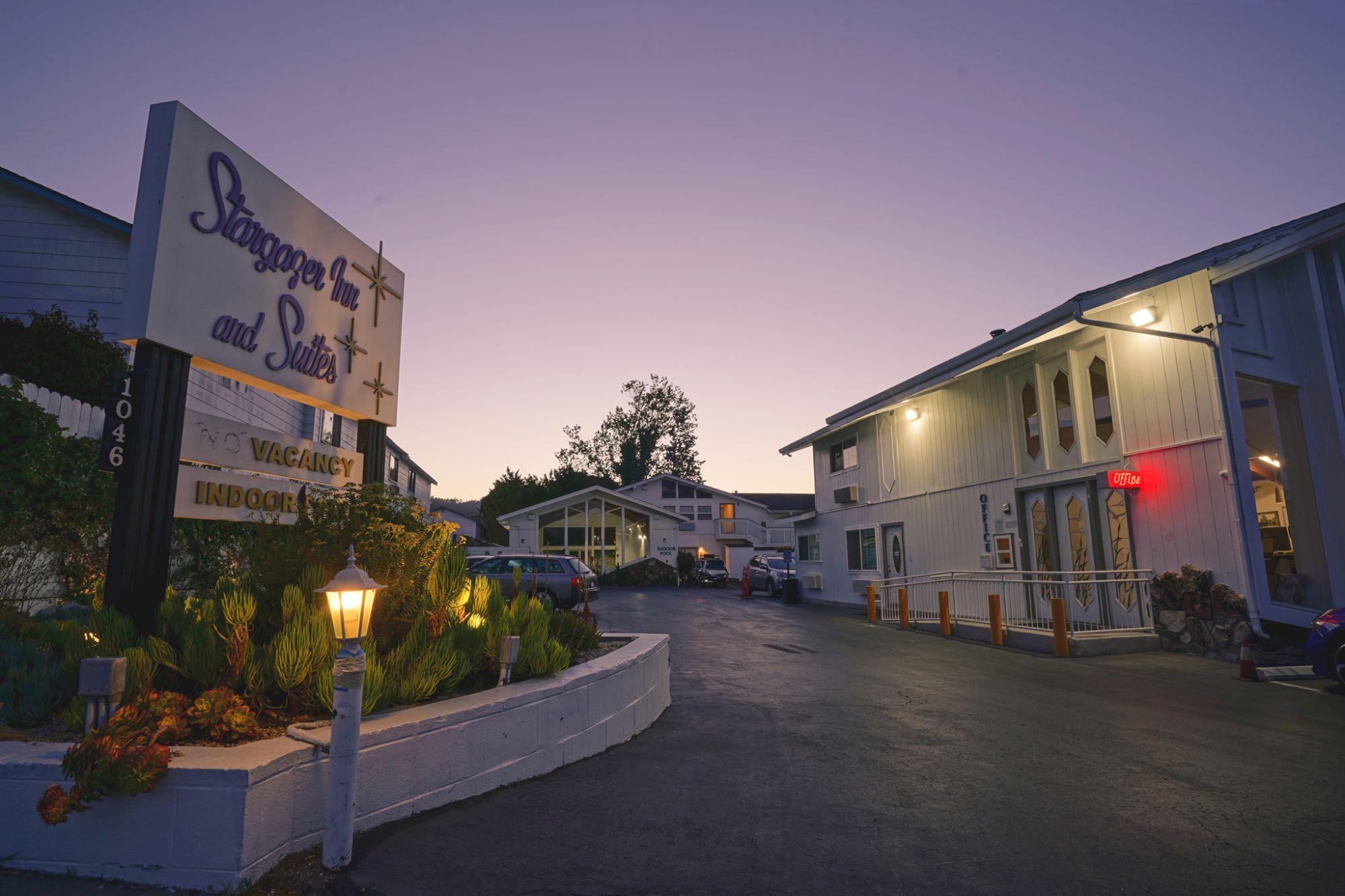 A motel entrance at dusk with a sign advertising rooms and suites, a driveway, and two-story buildings lit softly along the quiet road.